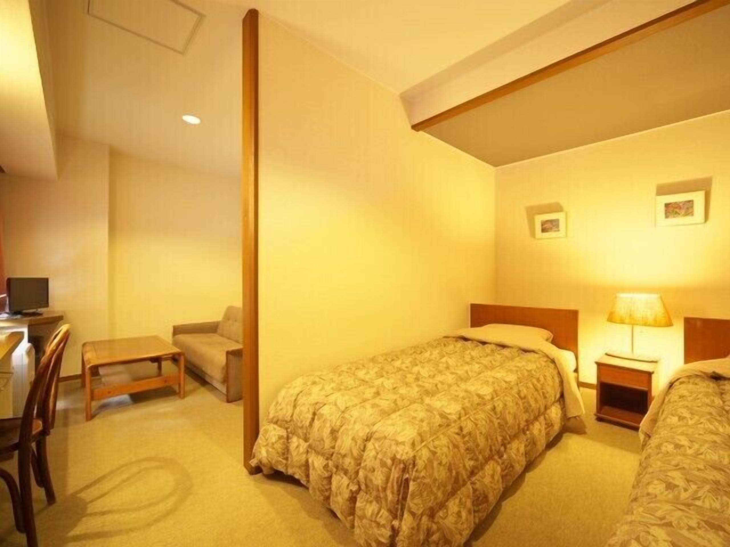 A hotel room with a single bed, a desk, a chair, and a lamp.