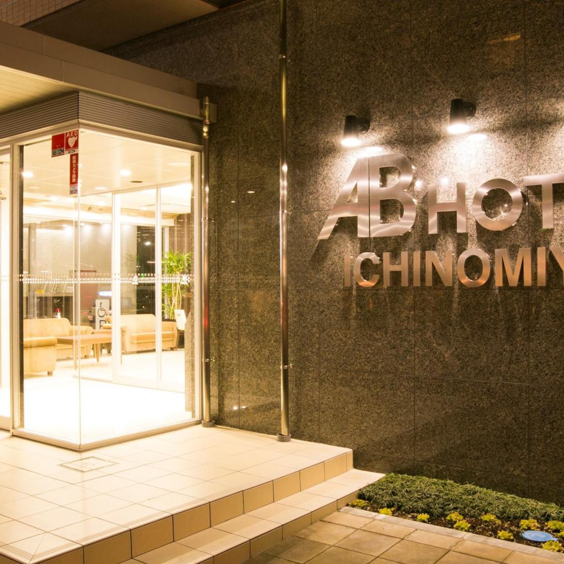 Ingresso di un hotel con porta in vetro, un cartello con su scritto 'ABHOT CHINOMI' e una piccola area giardino con piante.