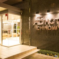 Entrada de hotel con puerta de vidrio, un letrero que dice 'ABHOT CHINOMI' y un pequeño jardín con plantas.