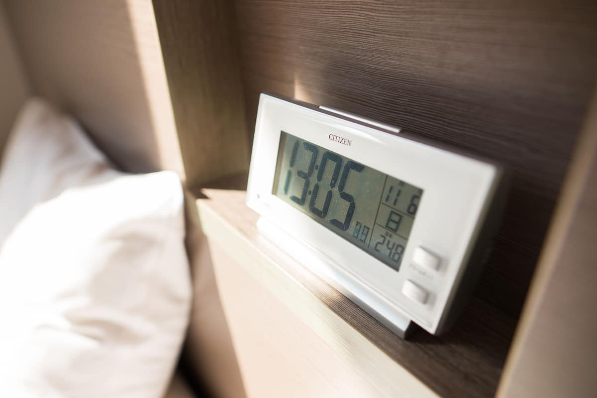 Een digitale klok toont 13:05 op een nachtkastje in een hotelkamer.