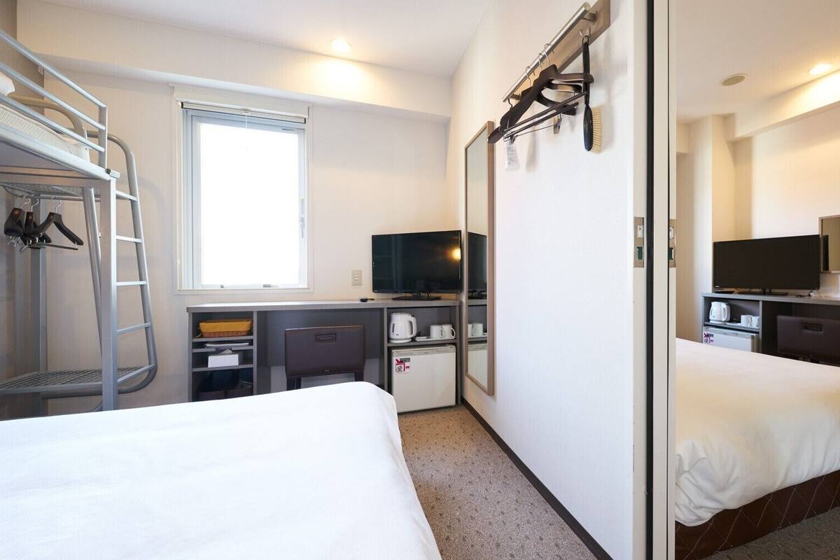 Een hotelkamer met een stapelbed, een bureau met een computer, een televisie, een kleine koelkast en een spiegel.