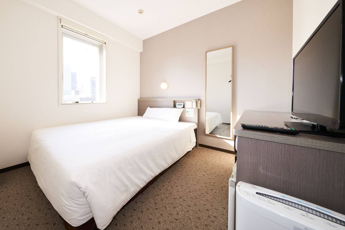 Een hotelkamer met een enkel bed, een televisie, een bureau met een computer, een spiegel en een raam met luxaflex.