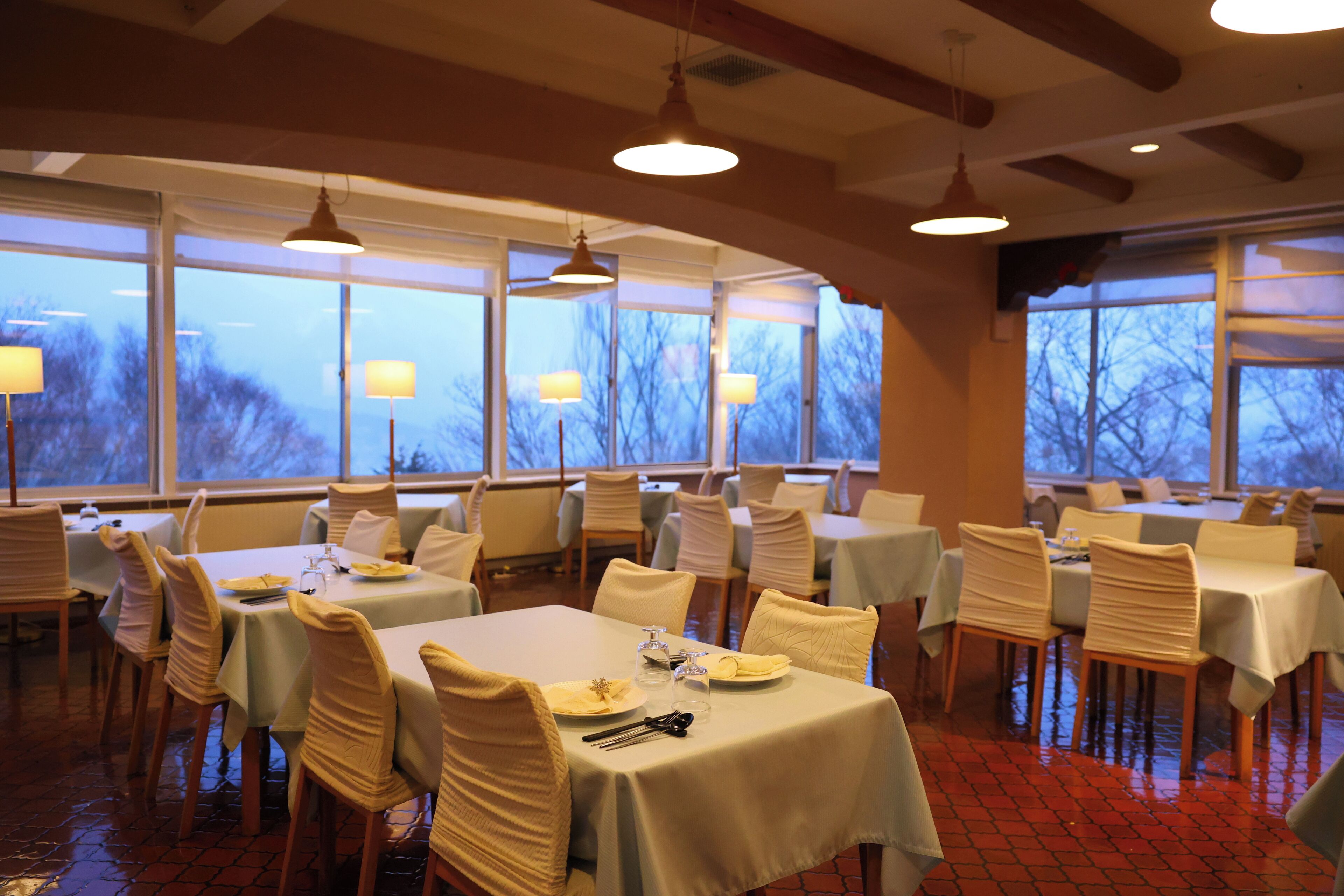A restaurant with tables set for dining, large windows, and hanging pendant lights.