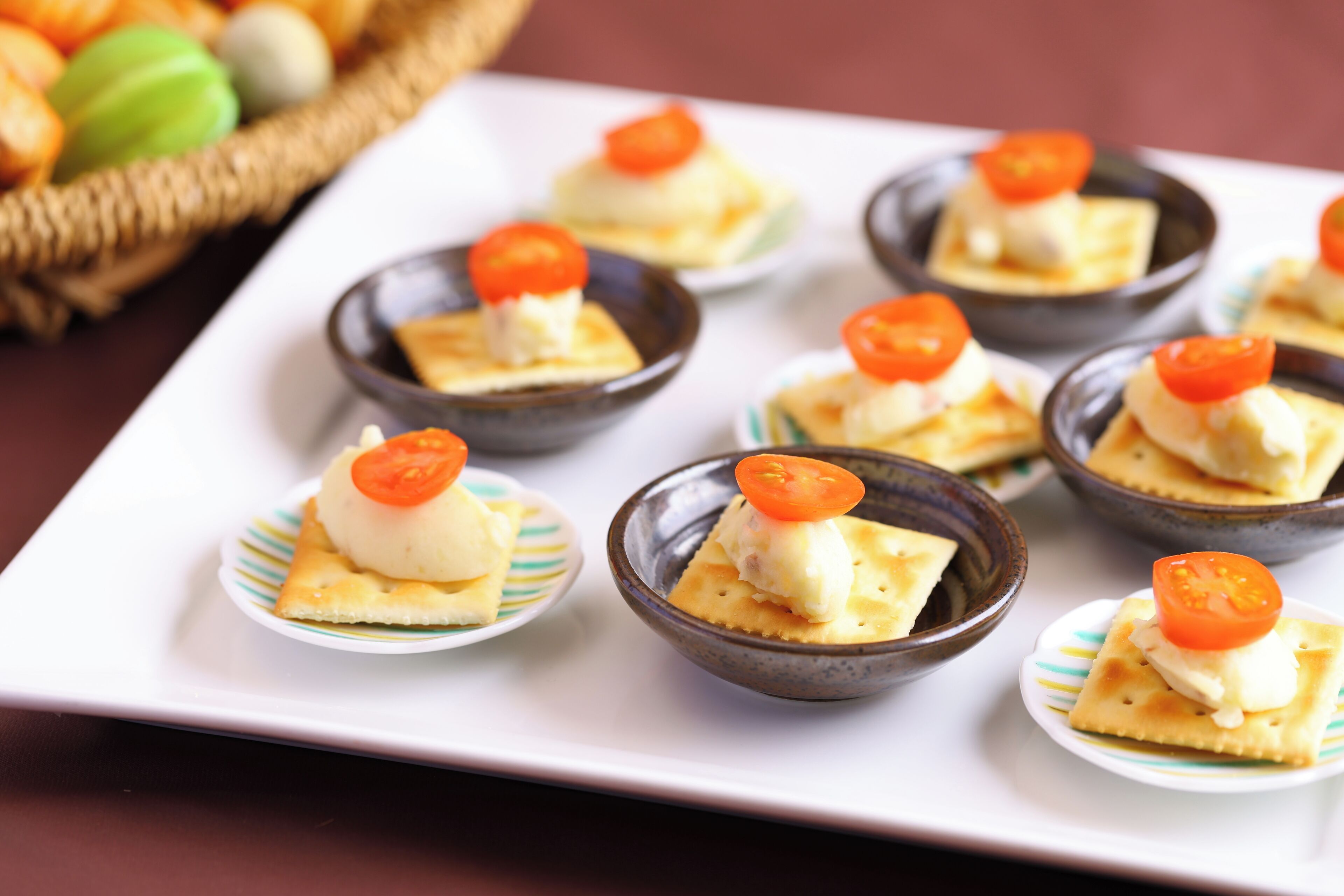 A white rectangular plate with small bowls containing a white creamy substance topped with a cherry tomato, placed on a striped paper plate.