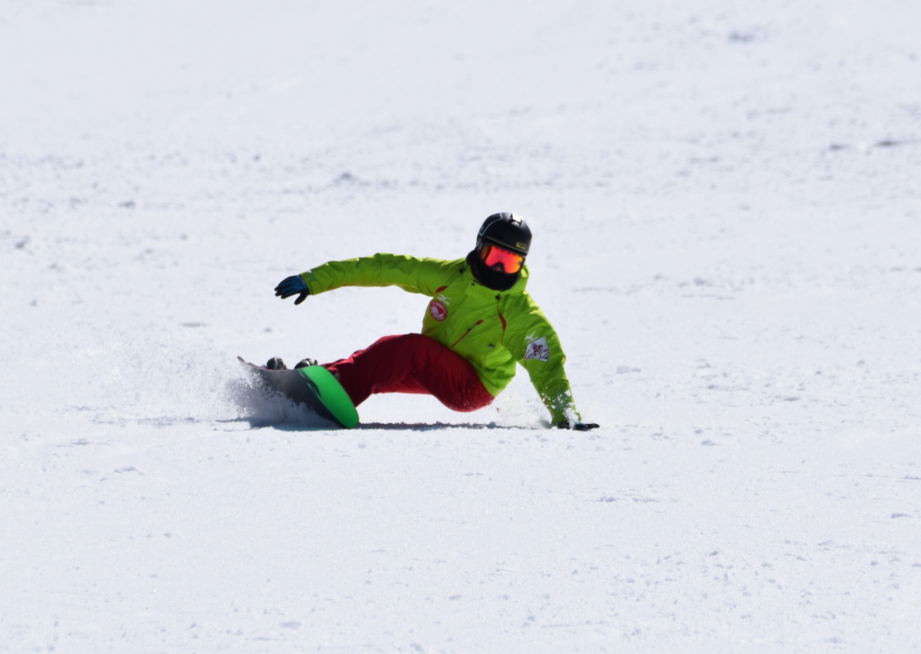 Ein Snowboarder in einer leuchtend grünen Jacke und roten Hose macht eine Schwungfahrt auf einem verschneiten Hang.