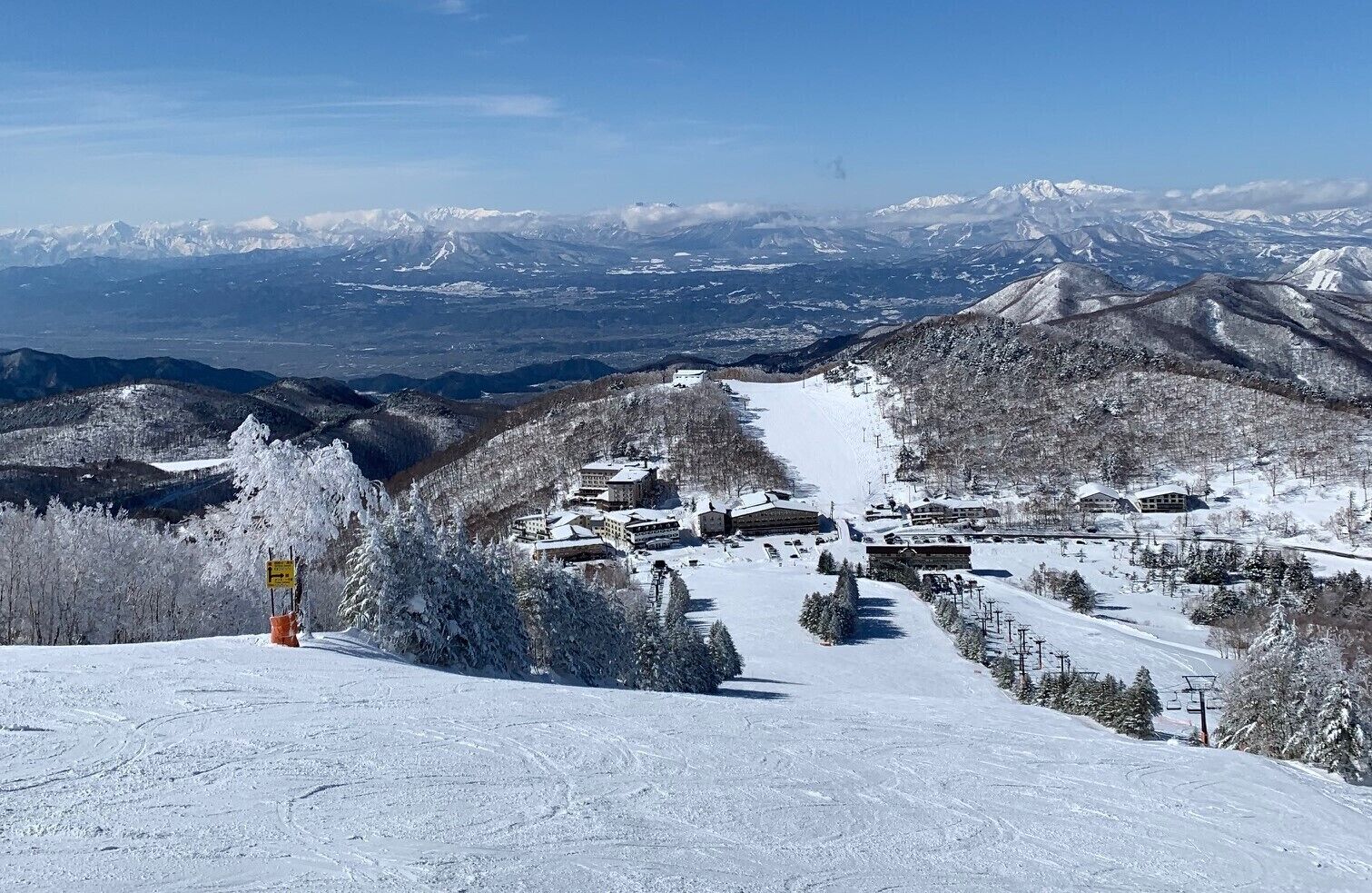 A snow-covered mountain resort with multiple buildings, ski slopes, and a clear view of distant snow-capped peaks.