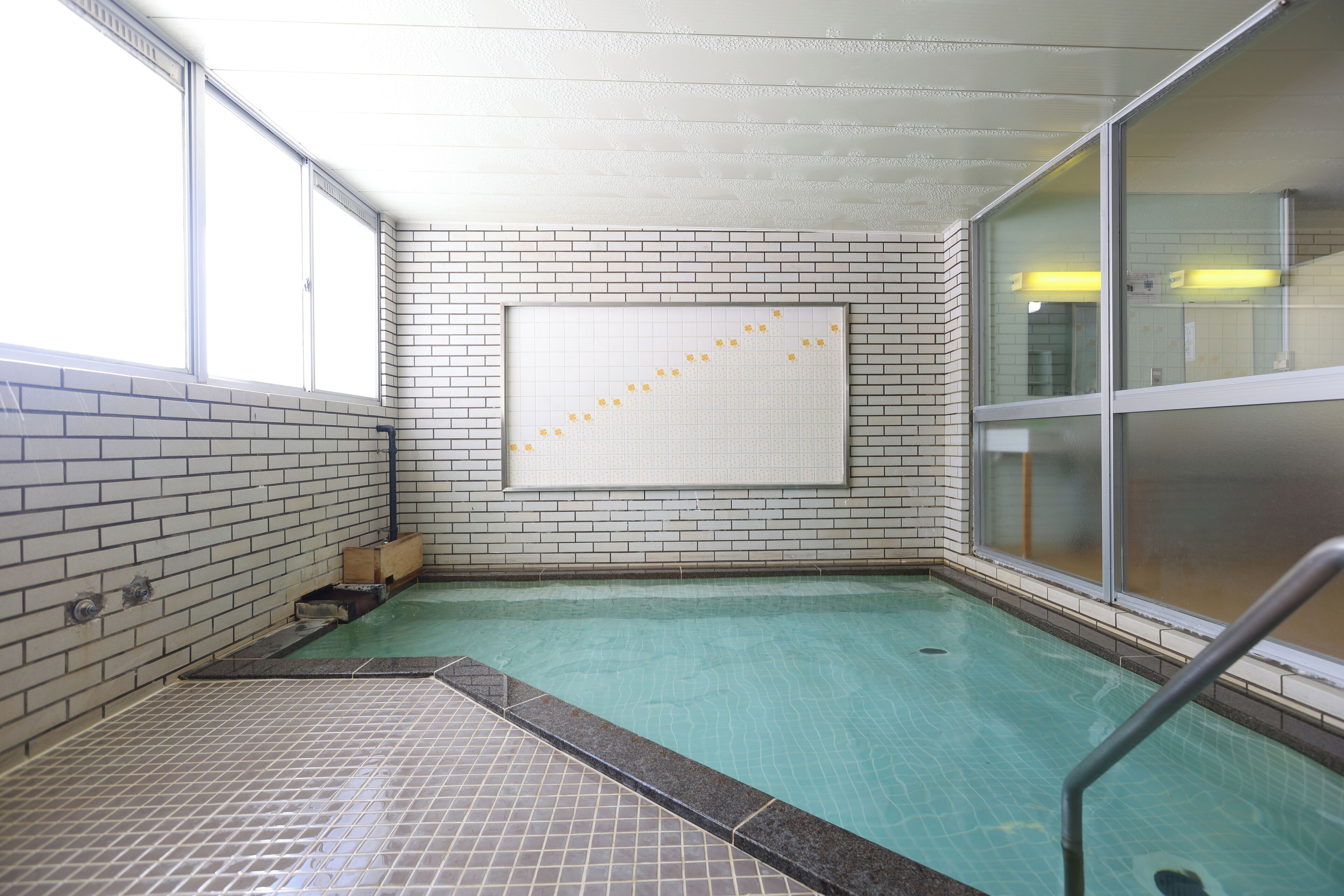 A swimming pool with tiled walls and a tiled floor, a window with blinds, and a metal handrail.