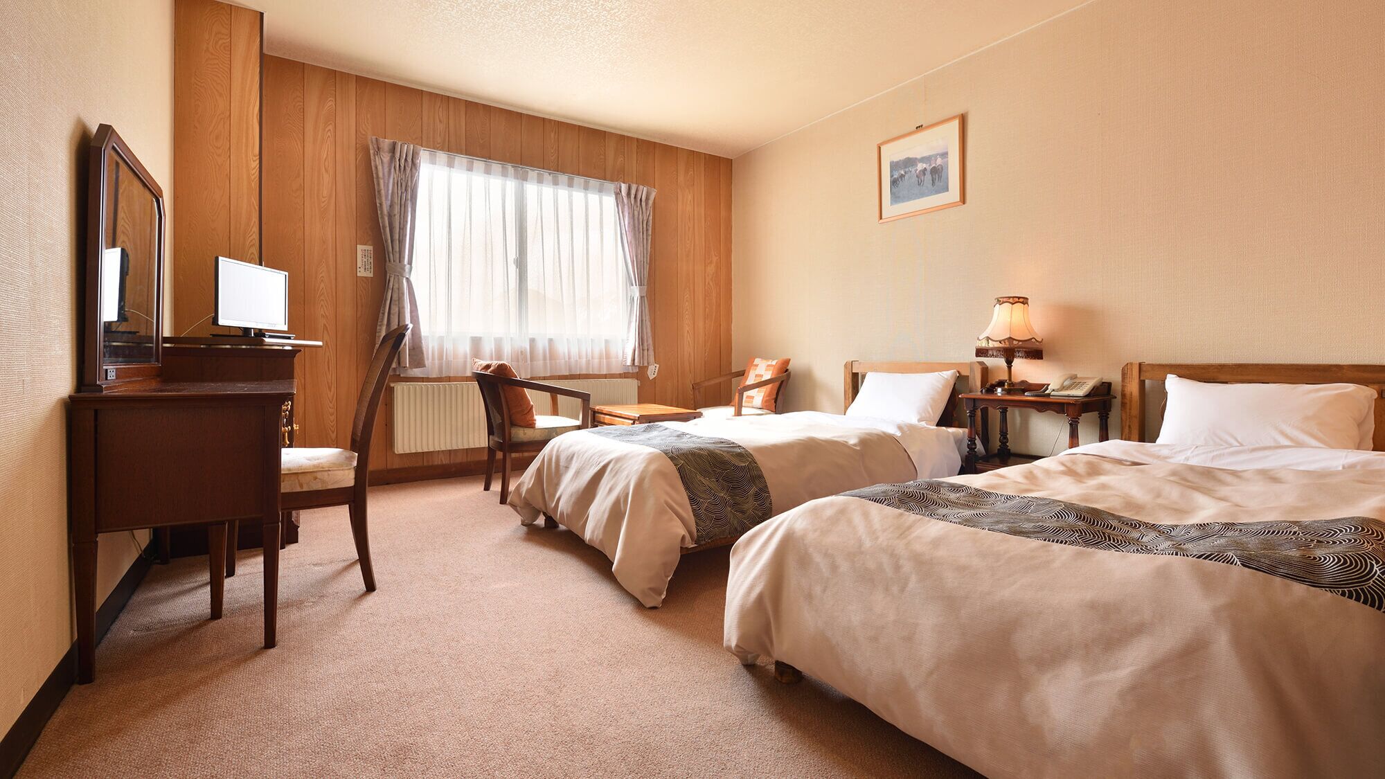 Ett hotellrum med två sängar, ett skrivbord, en stol, en lampa och ett fönster med gardiner.