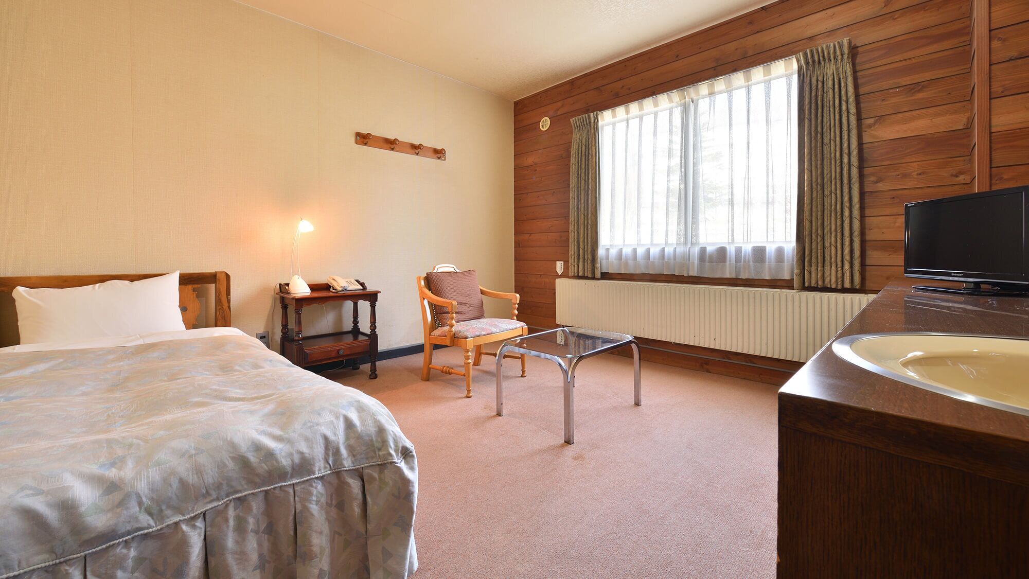 Ett hotellrum med en säng, en stol, ett skrivbord, en tv och ett fönster med gardiner.