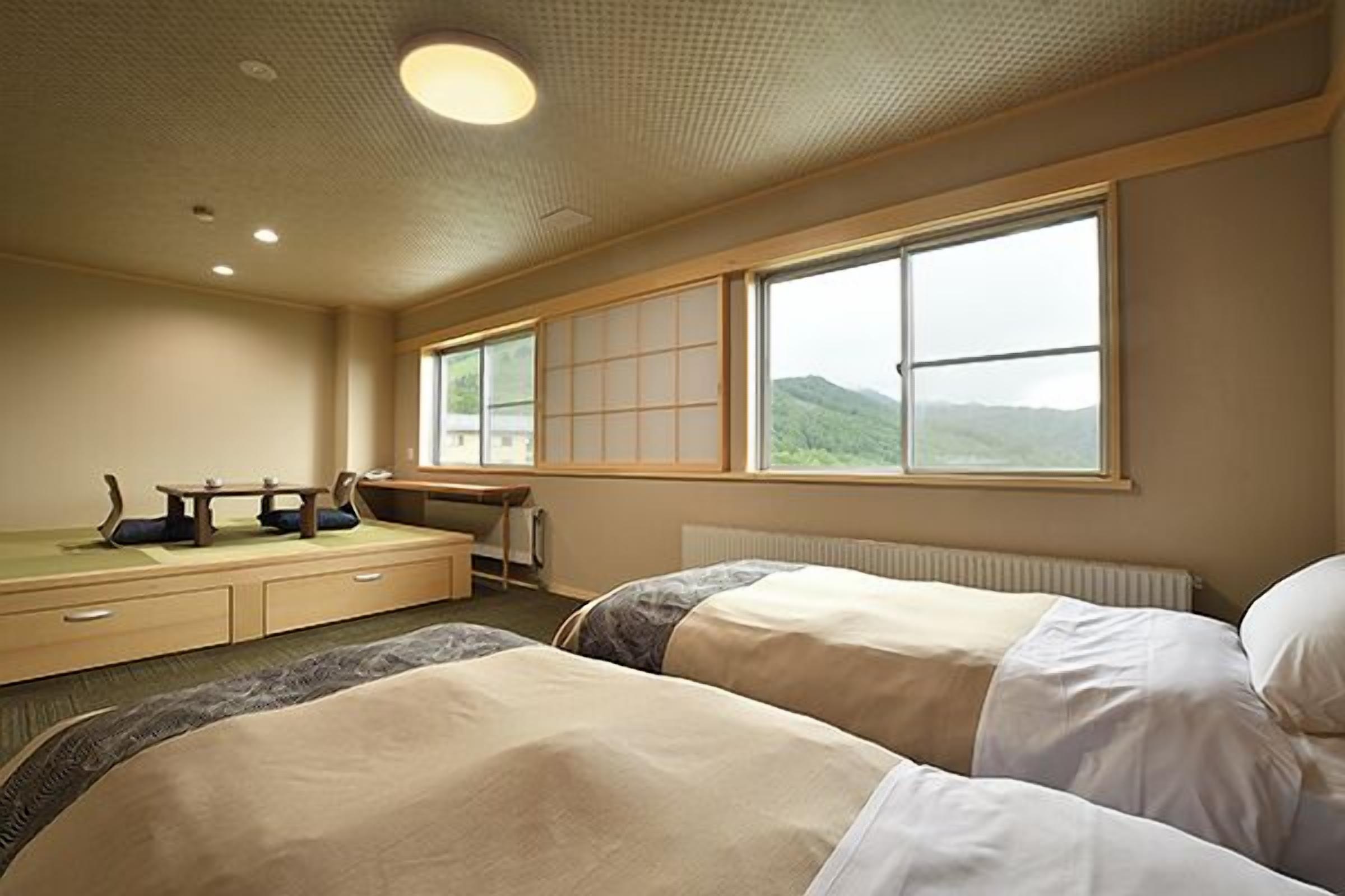 Une chambre d’hôtel avec deux lits, un bureau, une chaise et une grande fenêtre donnant sur des collines.