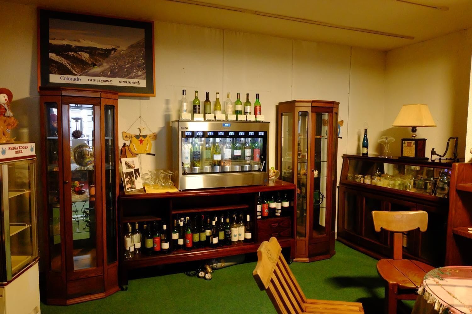 A bar area with a refrigerator stocked with bottles, a wooden chair, and a table with a lamp.