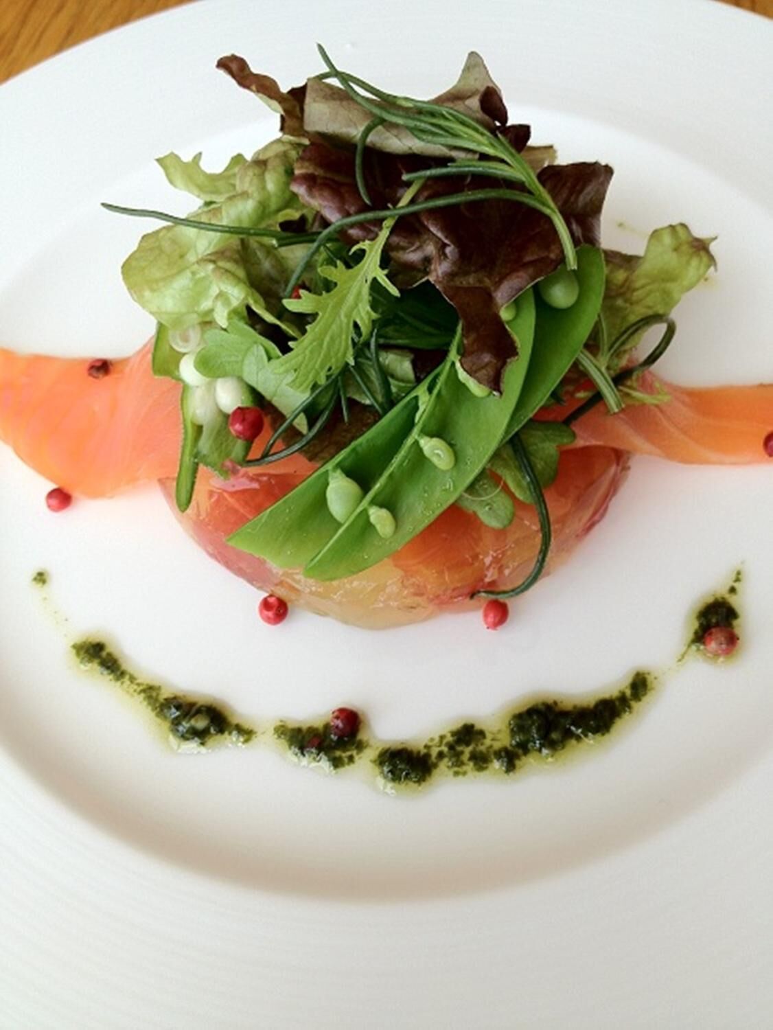 A plate with a dish of salmon, greens, and peas, garnished with red peppercorns and a green sauce.