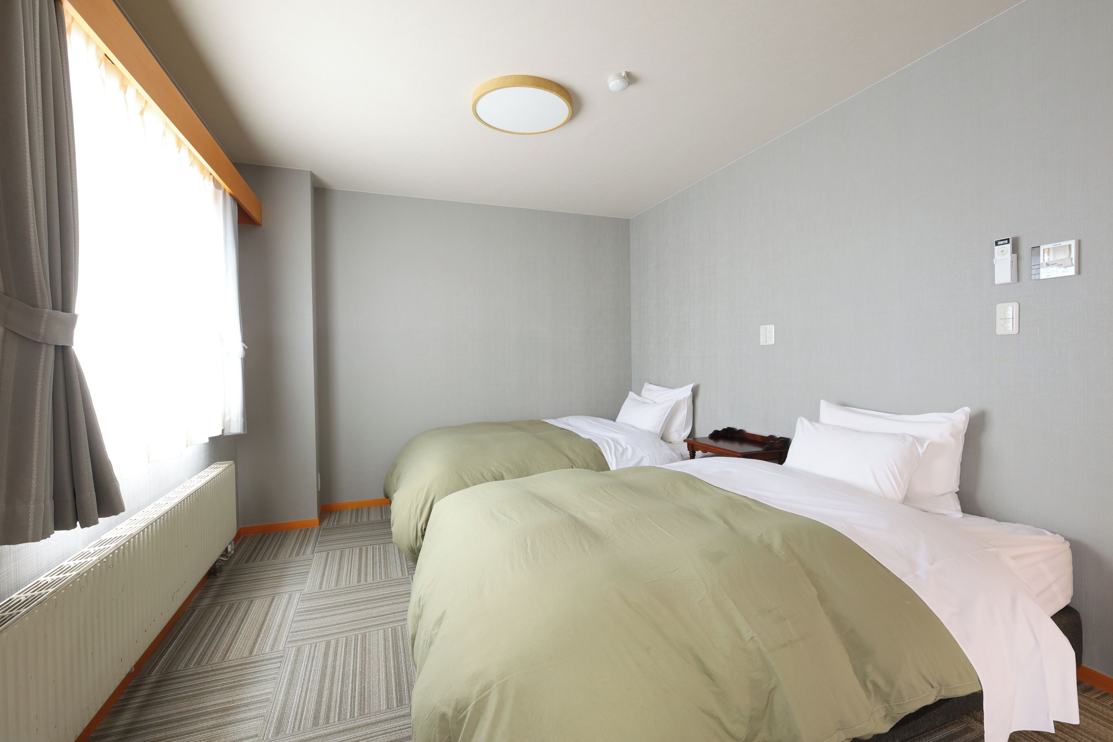 Ein Hotelzimmer mit zwei Betten, einem Heizkörper, einem Fenster mit Vorhängen und einer Deckenleuchte.