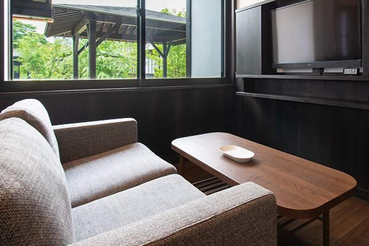 A modern room with a sofa, a small table, a TV mounted on the wall, and a window with a view of greenery.