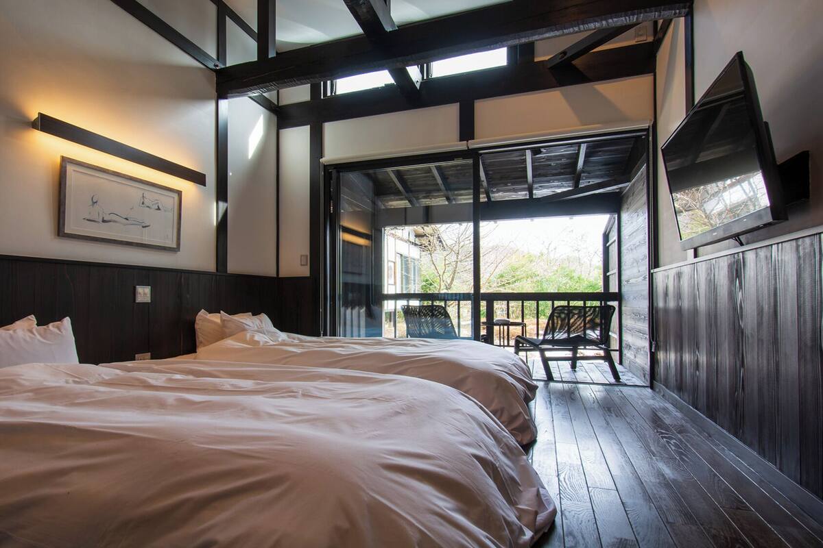A room with two beds, a framed artwork on the wall, and a view through an open door.