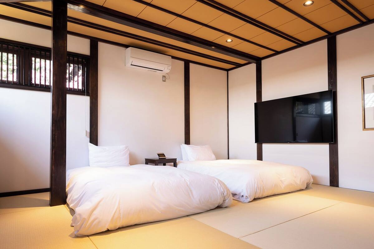 A traditional Japanese-style room with tatami flooring, two beds with white bedding, a mounted air conditioner, a small bedside table with a lamp, and a TV on the wall.