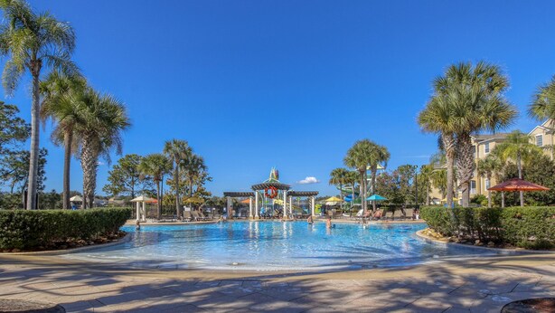 A large outdoor swimming pool surrounded by palm trees and shaded areas, with a pavilion in the center. - Lovely 5 bedroom pool and spa home with fantastic cinema in Windsor Hills Resort (Kissimmee)