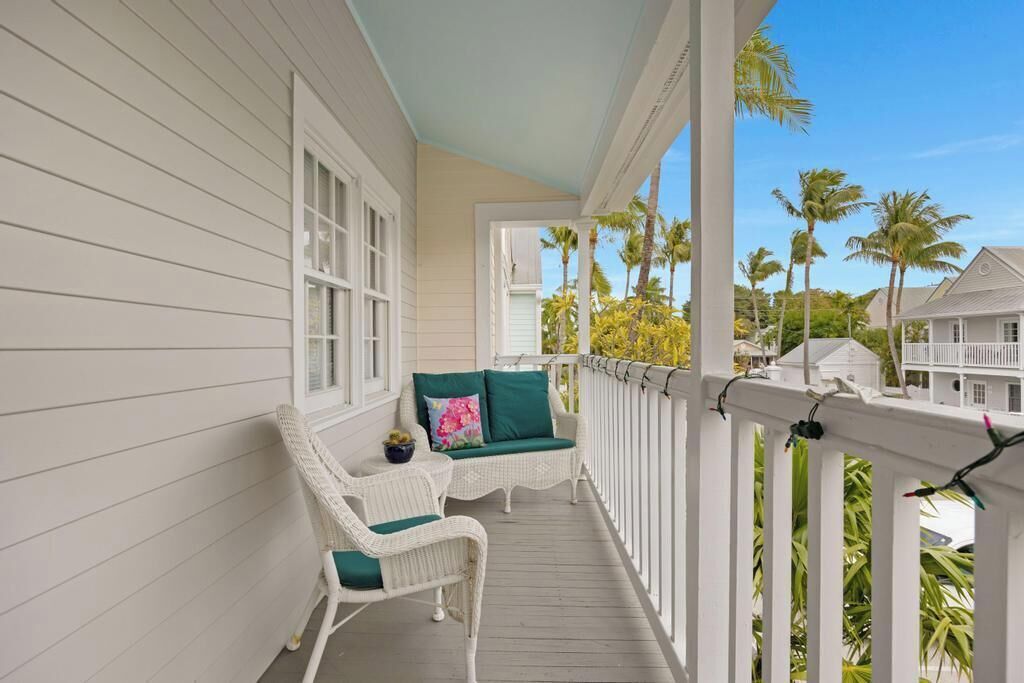 Un porche cubierto con muebles de mimbre blanco y cojines verdes, con vistas a una zona residencial con palmeras.