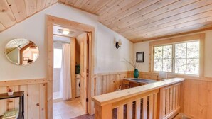 Interior de una cabaña de madera con techo de madera, un espejo redondo, una mesita, un ventanal y una puerta que da a otra habitación.