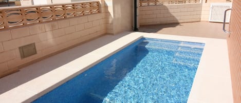 En rektangulär swimmingpool med klart, blått vatten, omgiven av beige kakelväggar och ett dekorativt trästaket.