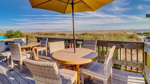 Eine Holzterrasse mit Korbmöbeln und einem gelben Sonnenschirm mit Blick auf einen Strand.
