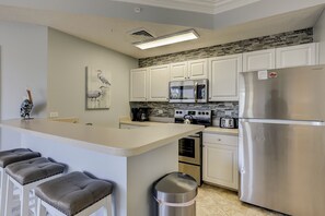A kitchen with white cabinets, a stainless steel refrigerator, a microwave, a stove, and a sink. There are bar stools and a trash can.