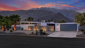 Une maison moderne à deux étages avec un garage, entourée d’une végétation désertique et avec des montagnes en arrière-plan.
