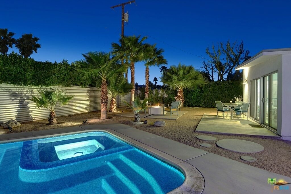 A backyard with a swimming pool, palm trees, a patio with outdoor furniture, and a modern house.