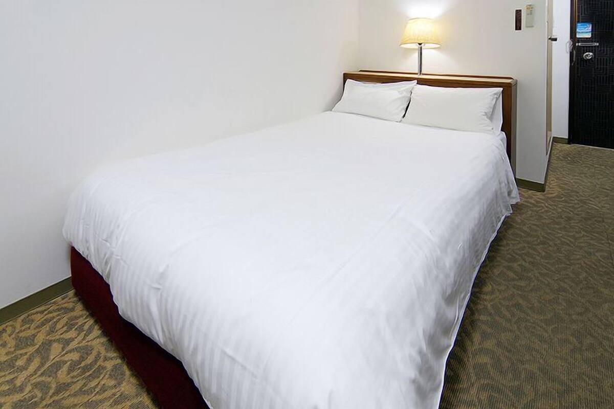 Una cama individual con ropa blanca, un cabecero de madera, una lámpara de pared y suelo alfombrado.
