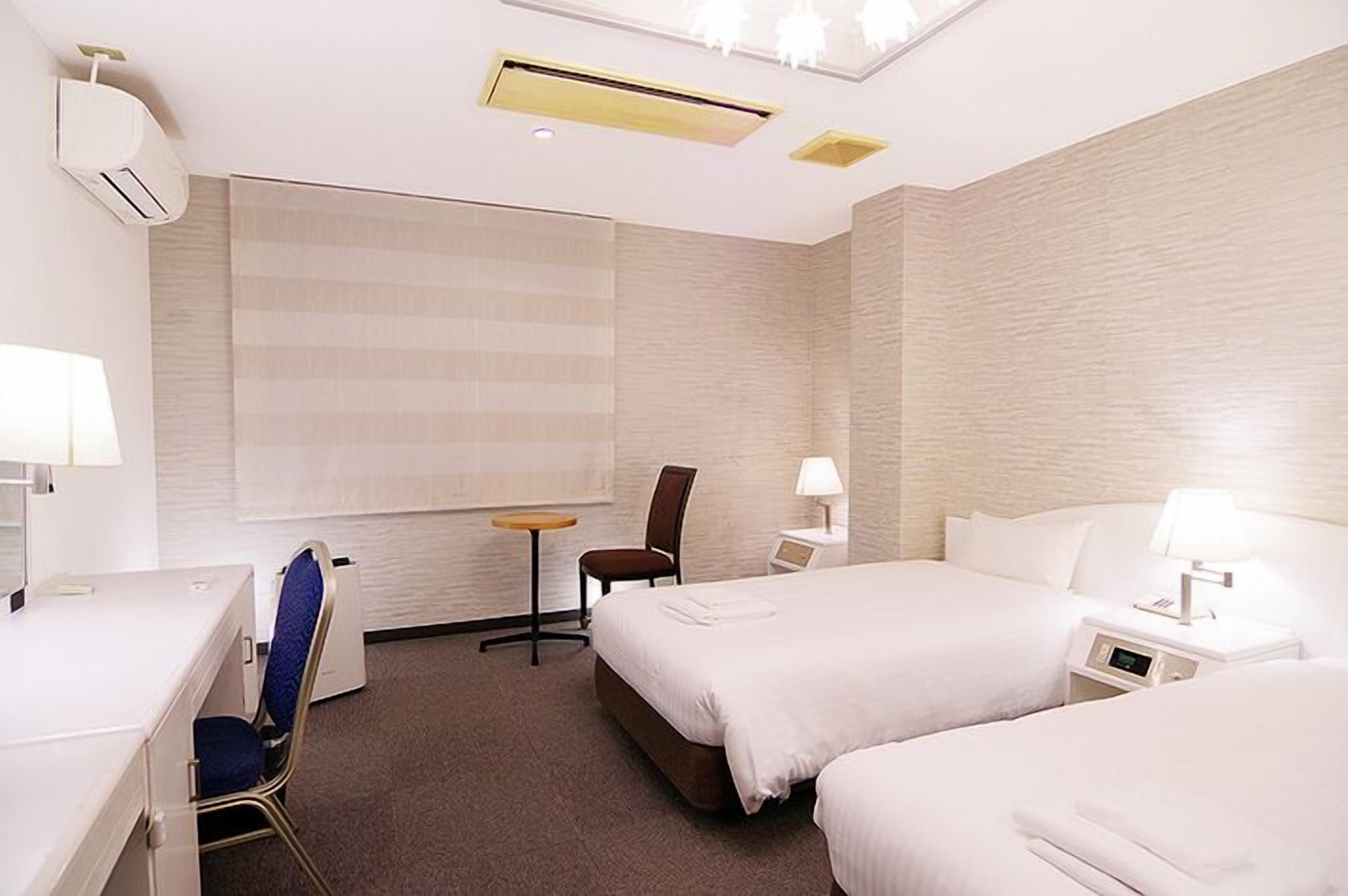 Habitación de hotel con dos camas, un escritorio, una silla, una mesita y un aire acondicionado de pared.