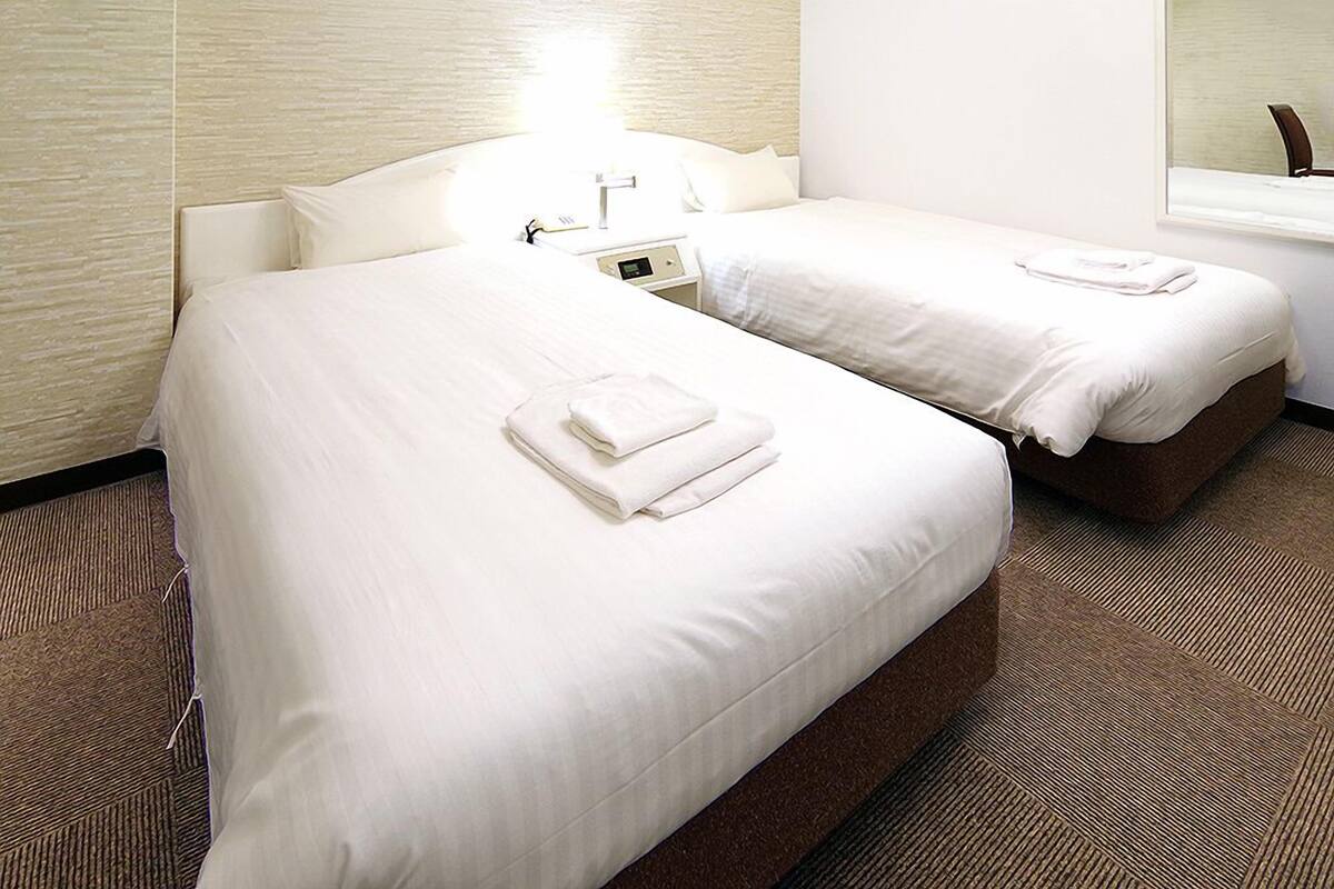 Una habitación de hotel con dos camas, una mesita de noche con lámpara y una ventana.