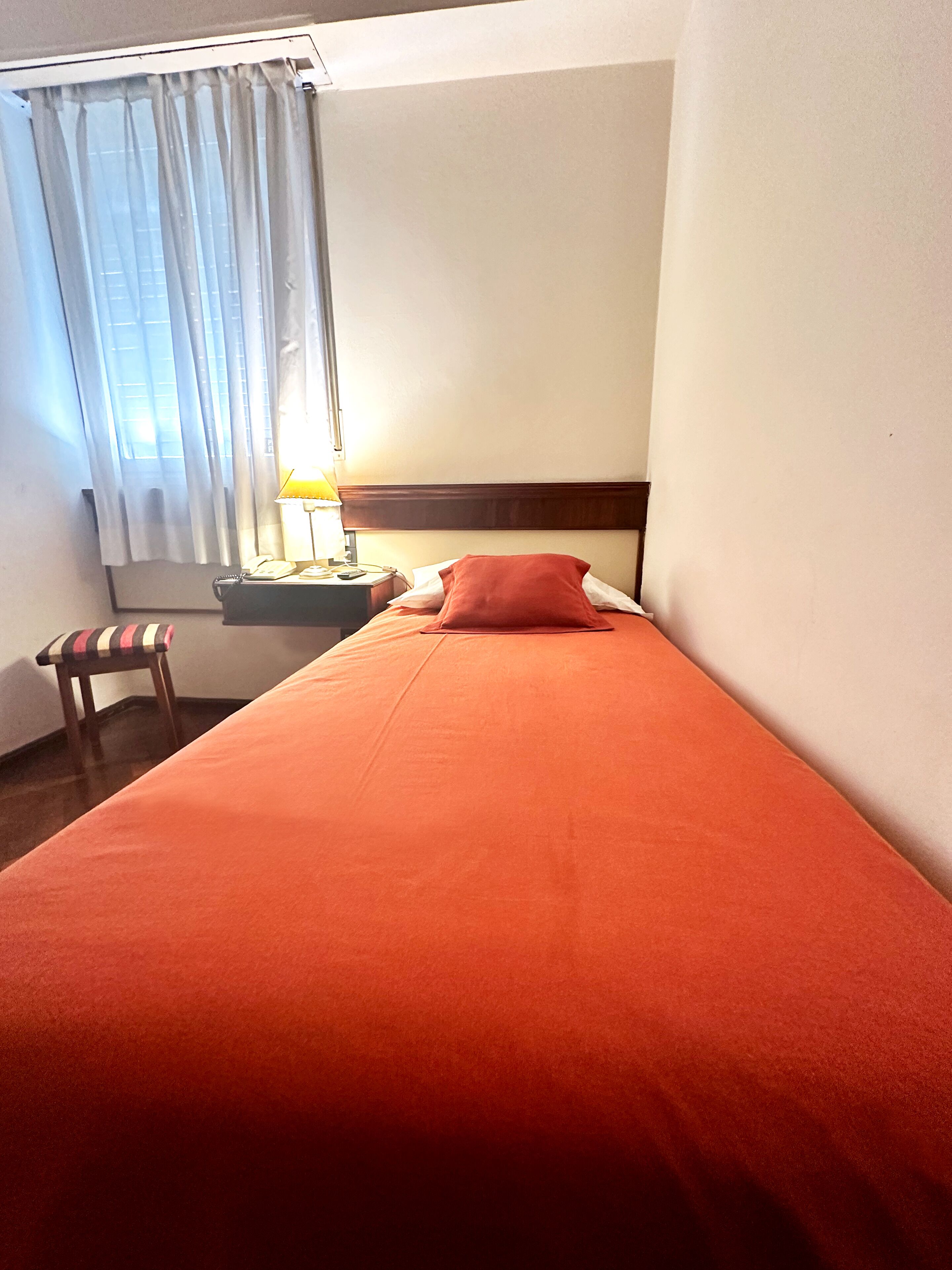 Una cama individual con una manta roja, un cabecero de madera, una mesita de noche con una lámpara, un teléfono y una silla con cojín a rayas.