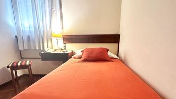 Un lit simple avec une couverture rouge, une tête de lit en bois, une table de chevet avec une lampe, un téléphone et une chaise avec un coussin à rayures.