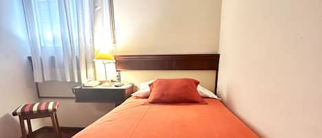 Una cama individual con una manta roja, un cabecero de madera, una mesita de noche con una lámpara, un teléfono y una silla con cojín a rayas.