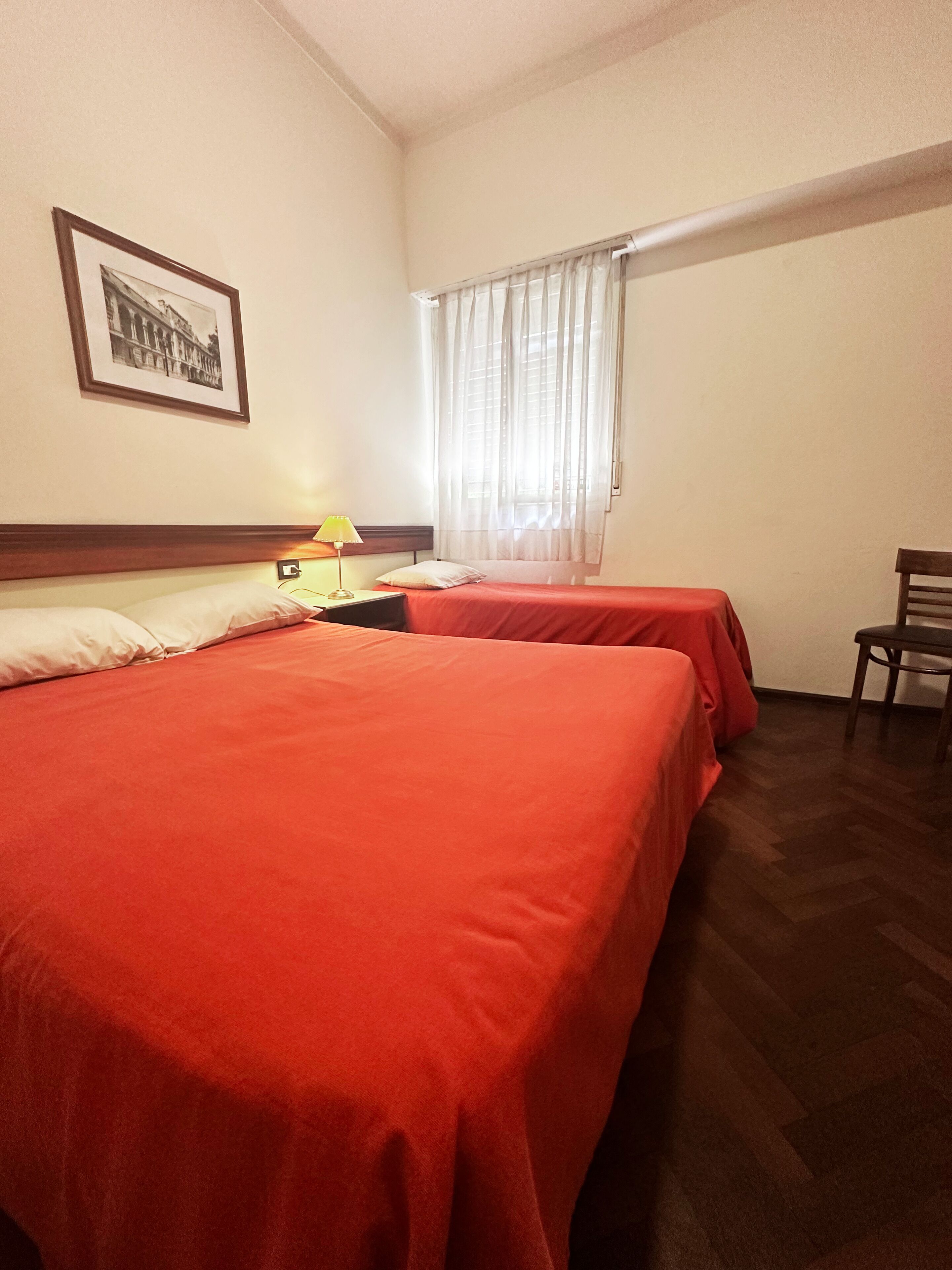 A hotel room with two beds, a desk, a chair, a lamp, and a window with curtains.