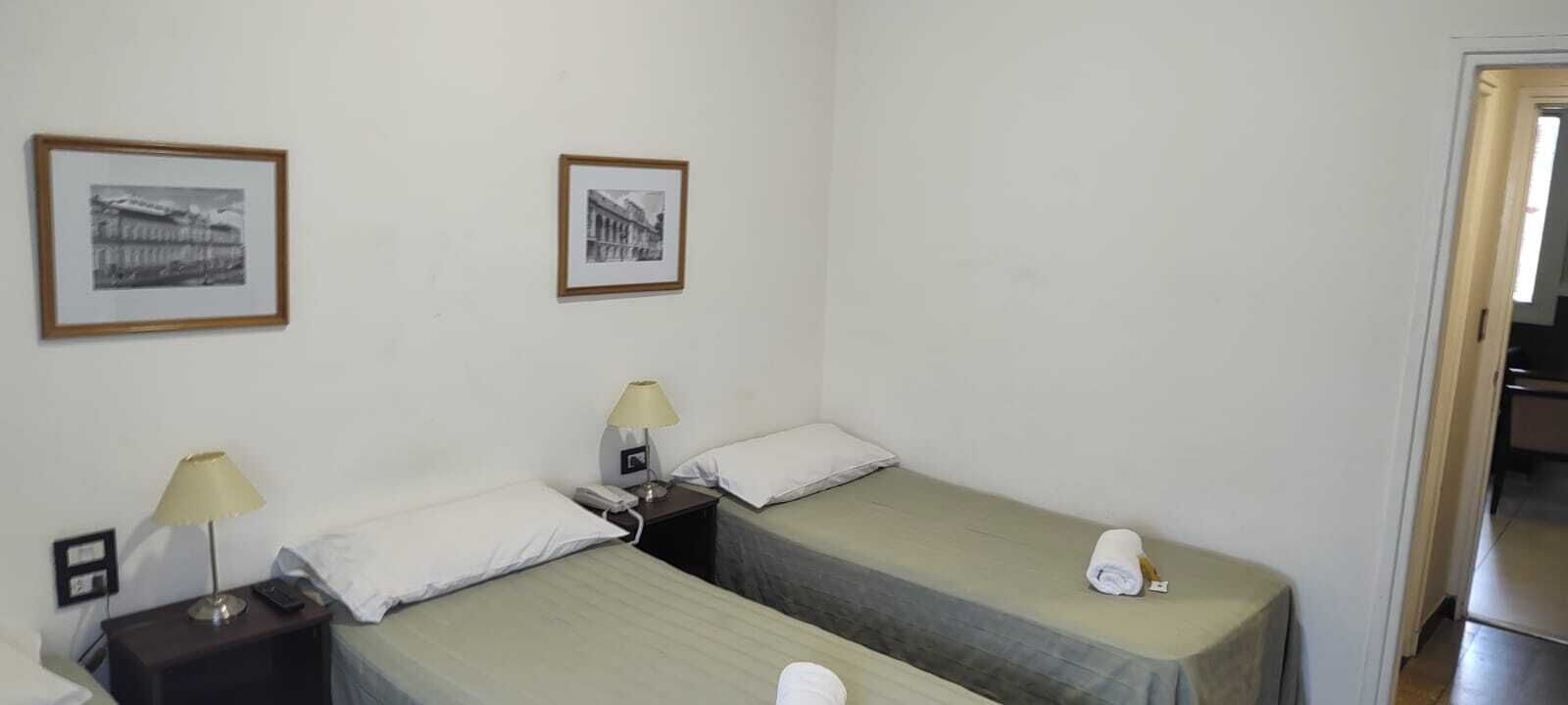 A simple room with two single beds, each with a nightstand, lamp, and a telephone. There are two framed pictures on the wall.