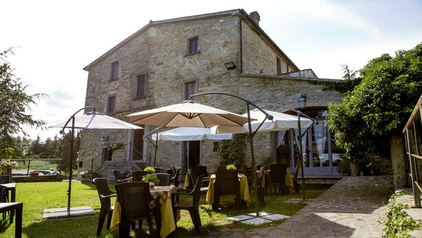 An outdoor patio with tables and chairs, a stone building in the background, and a pathway leading to the entrance. - Country House Santa Felicita la Paterna (Città di Castello)