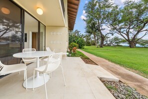 A patio with a white table and chairs, a glass door, and a view of a grassy area with trees. - Waiohuli Beach Hale #D-126 Deluxe Oceanfront, Best Location in Complex! (Kihei)
