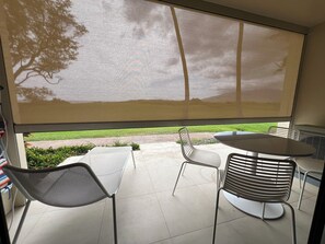 Un patio con mesa y sillas, un ventanal con mosquitero y vistas a un césped y árboles.