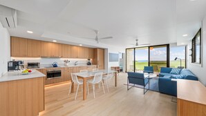 A modern kitchen with wooden cabinets, a dining area with white chairs, and a living room with a blue sofa and a view of the outdoors.