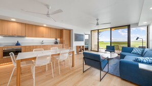 A modern kitchen with wooden cabinets, a dining area with white chairs, a blue sofa, and large windows offering a view of the outdoors.