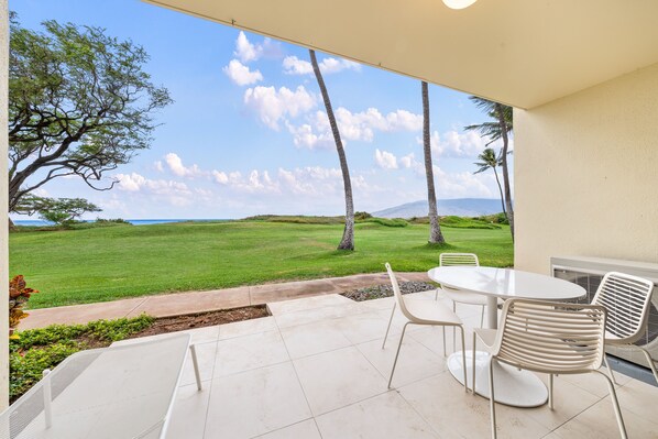 A patio with a table and chairs, overlooking a grassy area with trees and a distant view of mountains. - Waiohuli Beach Hale #D-126 Deluxe Oceanfront, Best Location in Complex! (Kihei)
