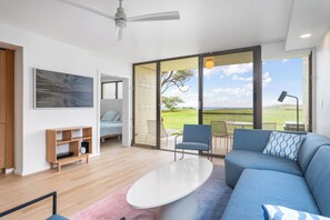 Un salon moderne avec un grand canapé bleu, une table basse blanche et une bibliothèque en bois. Une porte-fenêtre coulissante donne sur un patio extérieur d’où l’on peut admirer le ciel et un arbre.