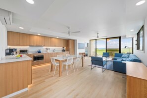 Une cuisine moderne avec des meubles en bois, un coin repas avec des chaises blanches et un salon doté d’un canapé bleu et offrant une vue sur l’extérieur.