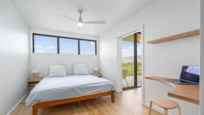 A bedroom with a bed, a desk with a laptop, a chair, a ceiling fan, and a view of the outdoors.