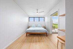 Une chambre avec un plancher en bois, un lit recouvert d’une courtepointe blanche, deux tables de chevet avec des lampes, un ventilateur de plafond et une fenêtre donnant sur l’extérieur.