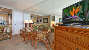 Een woonkamer met een eettafel, rieten stoelen, een televisie en een keuken op de achtergrond.