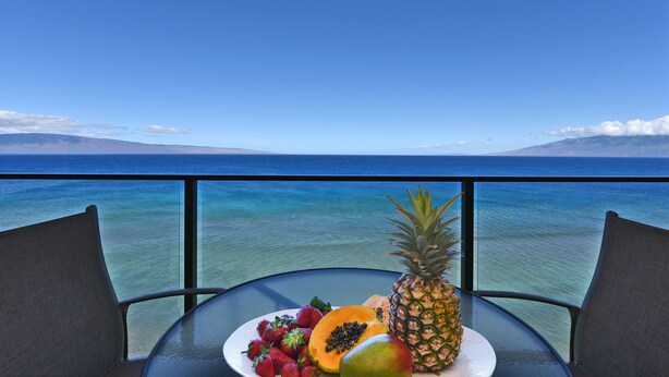 Een tafel met een fruitschaal en een stoel, met uitzicht op een heldere blauwe zee en verre bergen.