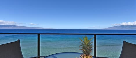 A table with a fruit platter and a chair, overlooking a clear blue sea and distant mountains.
