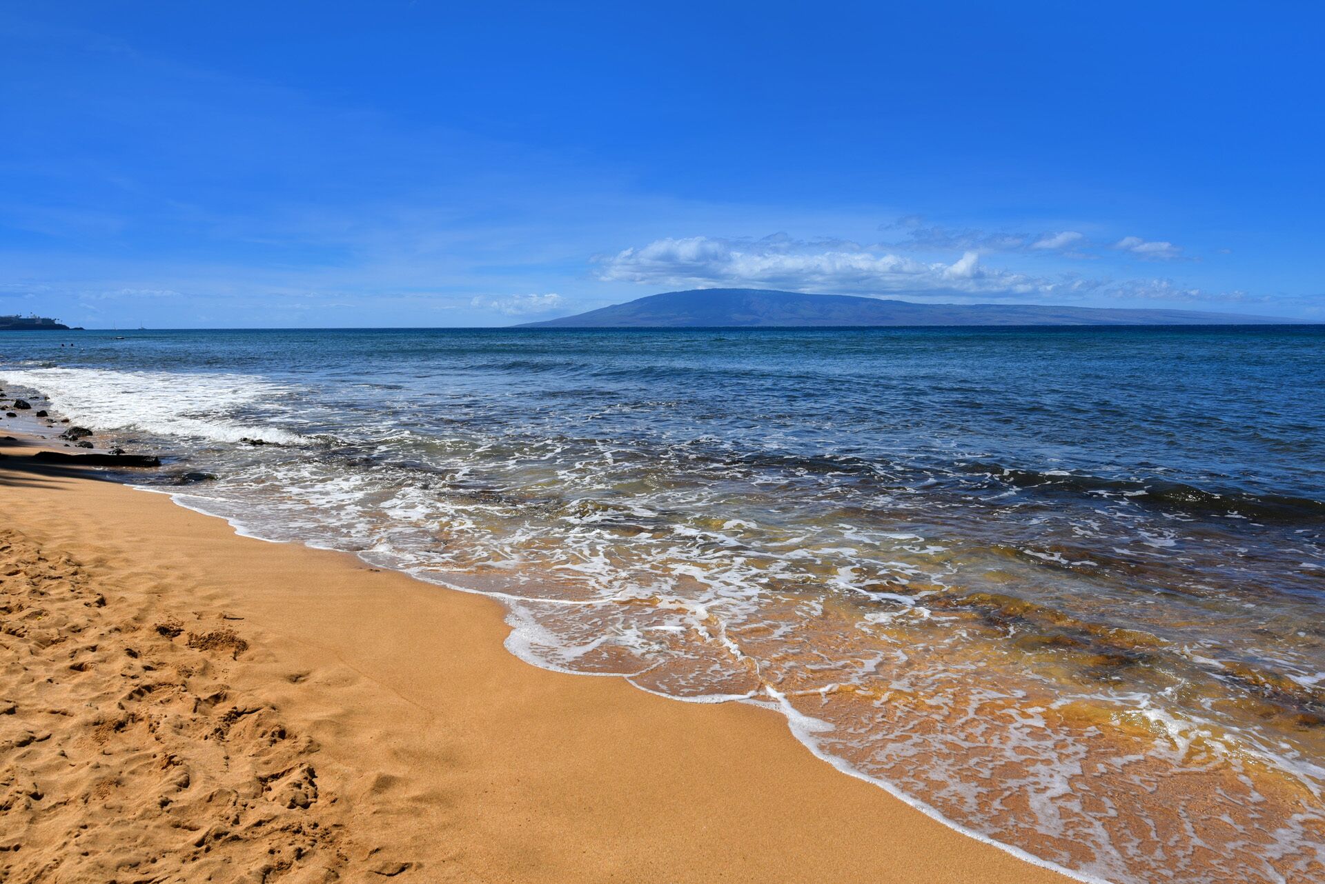 A beach with clear blue water, sandy shore, and a mountain in the distance.